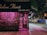 A florist shop named 'Chelsea Florist' with a dark green awning and a neon pink light illuminating the interior visible through the shop's window. The shop appears to be closed, with metal grates pulled over the entrance and window. The background shows a dimly lit city street at night, including a sidewalk, street signs, and various urban buildings.