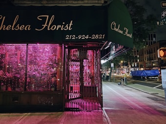 A florist shop named 'Chelsea Florist' with a dark green awning and a neon pink light illuminating the interior visible through the shop's window. The shop appears to be closed, with metal grates pulled over the entrance and window. The background shows a dimly lit city street at night, including a sidewalk, street signs, and various urban buildings.