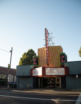 A vintage-style theater with an art deco marquee displaying movie titles. Neon lights outline the structure, creating a retro style. The marquee lists films including Barbie, Oppenheimer, and Mission Impossible. Bicycles are parked at the front, suggesting an urban setting.
