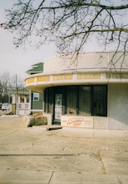 A small building with a curved, textured facade and a sign reading 'The Adventure Store' along with 'Parking Only' is situated along a quiet street. The building is partially shaded by a tree with barren branches. The architecture includes a combination of decorative stone work and barred windows with a vintage style, and a house with a porch can be seen in the background.