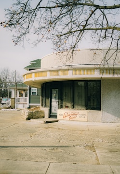 A small building with a curved, textured facade and a sign reading 'The Adventure Store' along with 'Parking Only' is situated along a quiet street. The building is partially shaded by a tree with barren branches. The architecture includes a combination of decorative stone work and barred windows with a vintage style, and a house with a porch can be seen in the background.