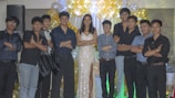 A group of people are standing together in a celebratory setting. A woman in a sparkling gown is in the center, surrounded by a group of men wearing formal attire. They are posing with arms crossed in front of a backdrop of gold and white balloons.