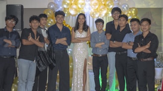 A group of people are standing together in a celebratory setting. A woman in a sparkling gown is in the center, surrounded by a group of men wearing formal attire. They are posing with arms crossed in front of a backdrop of gold and white balloons.