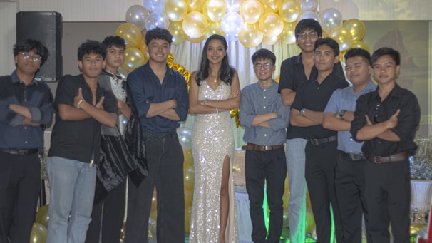 A group of people are standing together in a celebratory setting. A woman in a sparkling gown is in the center, surrounded by a group of men wearing formal attire. They are posing with arms crossed in front of a backdrop of gold and white balloons.