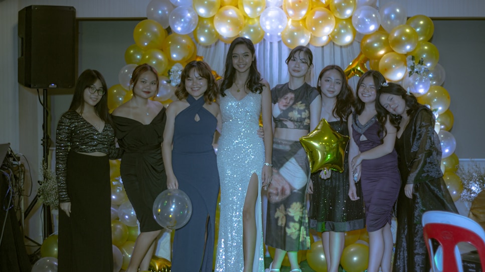 A group of eight women in formal attire stands in front of a backdrop made of gold and silver balloons. Some are holding balloons, and they are all smiling. The background features bright and festive decorations, suggesting a celebration or special event.