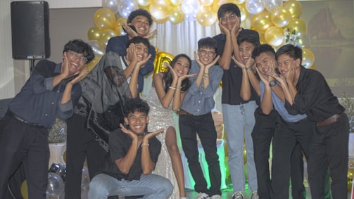 A group of people poses happily with their hands cupping their faces. They are dressed in casual to semi-formal attire, standing in front of a backdrop of white and gold balloons. The setting appears to be a celebration or party.