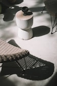 A minimalist outdoor setting with a small wooden stool placed on a light-colored surface. A dark ceramic cup rests on top of the stool. Surrounding the stool are woven rattan chairs casting intricate shadows on the ground due to the sunlight filtering through nearby foliage.