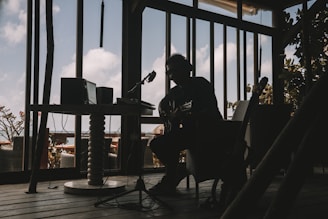 A silhouetted musician is sitting indoors with a guitar, surrounded by audio equipment. The room is dimly lit, creating strong contrasts between the dark interior and the bright sky visible through large windows. The décor suggests a relaxed, creative atmosphere.