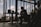 A silhouetted musician is sitting indoors with a guitar, surrounded by audio equipment. The room is dimly lit, creating strong contrasts between the dark interior and the bright sky visible through large windows. The décor suggests a relaxed, creative atmosphere.