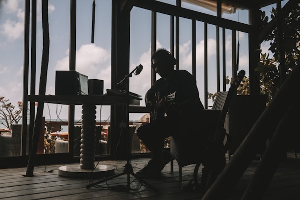 A silhouetted musician is sitting indoors with a guitar, surrounded by audio equipment. The room is dimly lit, creating strong contrasts between the dark interior and the bright sky visible through large windows. The décor suggests a relaxed, creative atmosphere.
