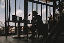 A silhouetted musician is sitting indoors with a guitar, surrounded by audio equipment. The room is dimly lit, creating strong contrasts between the dark interior and the bright sky visible through large windows. The d&eacute;cor suggests a relaxed, creative atmosphere.