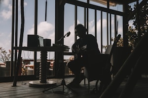A silhouetted musician is sitting indoors with a guitar, surrounded by audio equipment. The room is dimly lit, creating strong contrasts between the dark interior and the bright sky visible through large windows. The décor suggests a relaxed, creative atmosphere.
