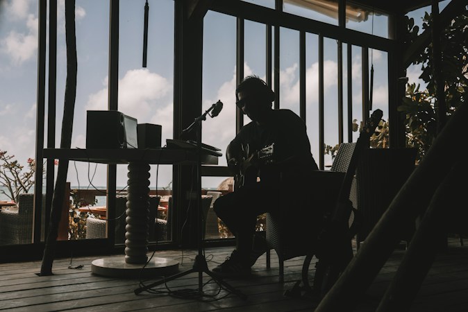 A silhouetted musician is sitting indoors with a guitar, surrounded by audio equipment. The room is dimly lit, creating strong contrasts between the dark interior and the bright sky visible through large windows. The d&eacute;cor suggests a relaxed, creative atmosphere.