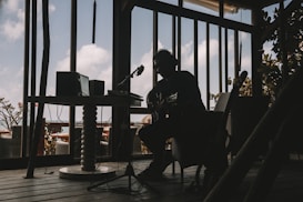 A silhouetted musician is sitting indoors with a guitar, surrounded by audio equipment. The room is dimly lit, creating strong contrasts between the dark interior and the bright sky visible through large windows. The décor suggests a relaxed, creative atmosphere.