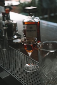 A cocktail glass filled with a dark beverage garnished with a stirring stick is placed on a bar counter. Nearby, a large bottle of liquor is prominently displayed along with other bar tools and bottles.