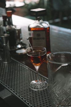 A cocktail glass filled with a dark beverage garnished with a stirring stick is placed on a bar counter. Nearby, a large bottle of liquor is prominently displayed along with other bar tools and bottles.