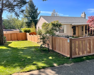 A quaint single-story house with a light-colored exterior and a gray shingle roof is surrounded by a wooden fence. The yard is well maintained with a lush green lawn and a few trees, including a tall evergreen and a tree with red leaves. The house is set under a clear blue sky, and sunlight bathes the entire scene.