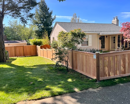A quaint single-story house with a light-colored exterior and a gray shingle roof is surrounded by a wooden fence. The yard is well maintained with a lush green lawn and a few trees, including a tall evergreen and a tree with red leaves. The house is set under a clear blue sky, and sunlight bathes the entire scene.