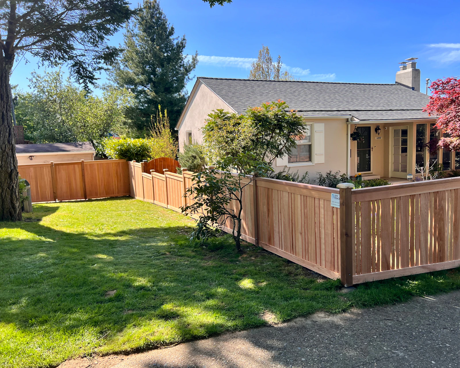 Single-story beige bungalow with a new cedar picket fence, small trees, and a tidy green lawn under a blue sky