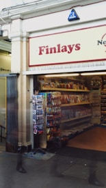 A small, cozy newsstand or convenience store named Finlays features a collection of magazines, newspapers, and various snacks on display. The entrance is open, revealing shelves stocked with printed materials and a rack with accessories or gifts. The store is located in a building with white walls and ornate decorative trim. There are blurry figures of people walking past the entrance, adding a sense of movement.