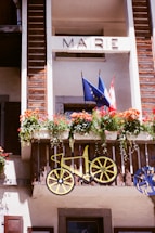 A balcony with wooden panels features a decorative bicycle motif and a floral arrangement with vibrant flowers. Two flags, one with European Union stars and the other likely belonging to a country, are displayed. The word 'MAIRIE' is written above.