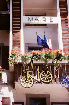 A balcony with wooden panels features a decorative bicycle motif and a floral arrangement with vibrant flowers. Two flags, one with European Union stars and the other likely belonging to a country, are displayed. The word 'MAIRIE' is written above.