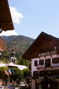 A picturesque alpine village scene featuring a traditional wooden building labeled 'Residence des Alpes' with an intricate roof design. In the background, a classic church with a steeple stands amidst lush green trees, set against a backdrop of a forested mountain. National flags are displayed in the foreground.
