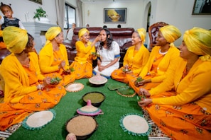 A group of women wearing vibrant yellow traditional attire and headscarves are sitting on a green carpet. They are engaged in a cultural or ceremonial activity, involving small baskets and bowls with green leaves and other items. One woman in the center is in white attire, and the group appears to be in a joyful and celebratory mood.