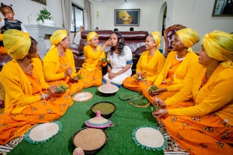 A group of women wearing vibrant yellow traditional attire and headscarves are sitting on a green carpet. They are engaged in a cultural or ceremonial activity, involving small baskets and bowls with green leaves and other items. One woman in the center is in white attire, and the group appears to be in a joyful and celebratory mood.