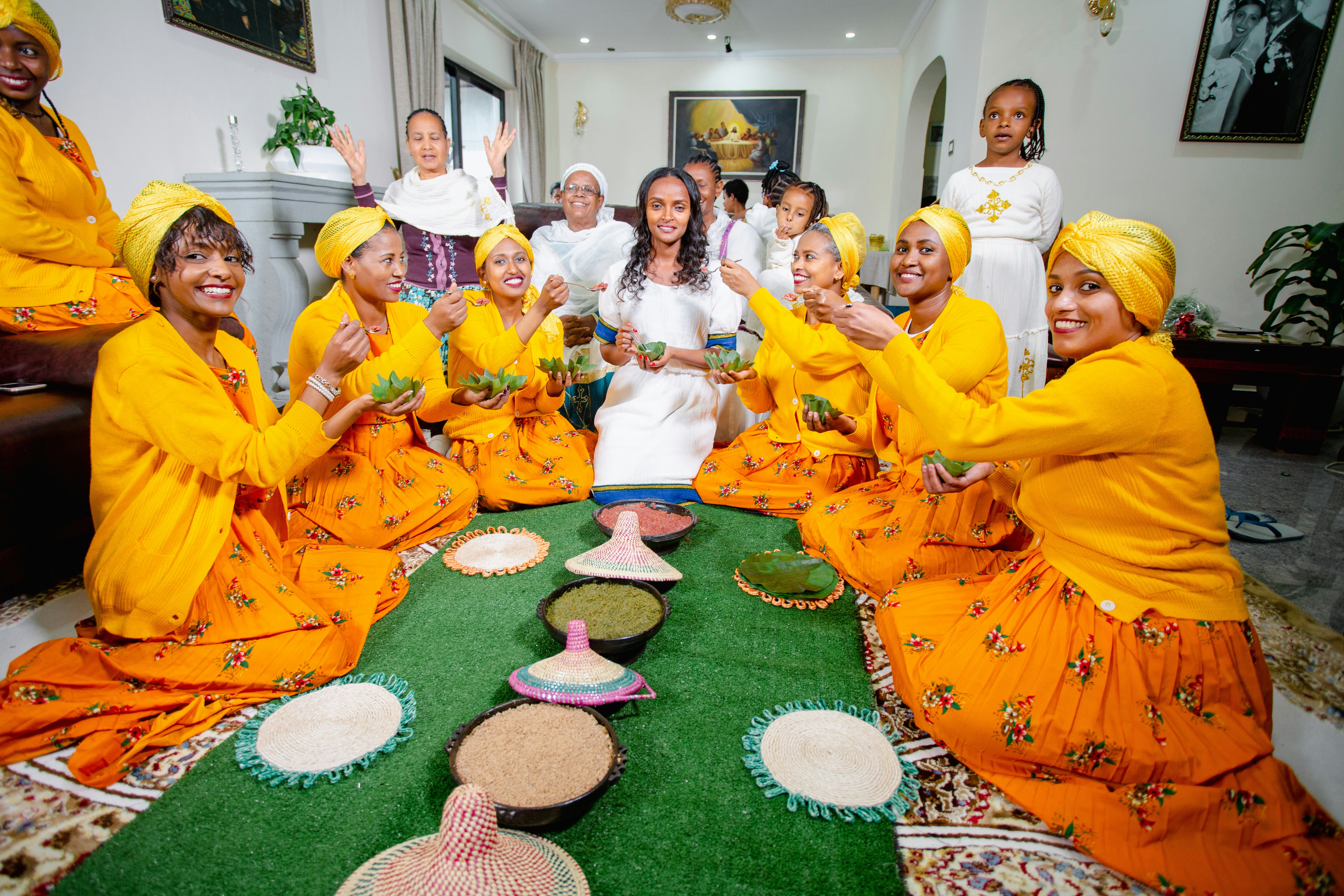 A group of people, predominantly women, dressed in brightly colored traditional attire are seated in a semi-circle around a green mat. They are holding green bowls and appear to be participating in a communal activity or celebration. Traditional woven baskets and plates filled with grains are placed on the mat. The room is well-lit and decorated with paintings and plants.