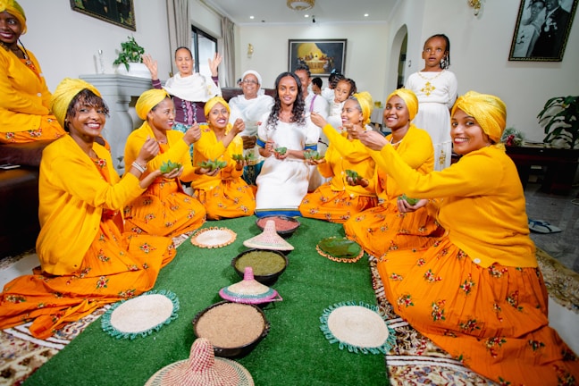 A group of people, predominantly women, dressed in brightly colored traditional attire are seated in a semi-circle around a green mat. They are holding green bowls and appear to be participating in a communal activity or celebration. Traditional woven baskets and plates filled with grains are placed on the mat. The room is well-lit and decorated with paintings and plants.