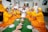 A group of people, predominantly women, dressed in brightly colored traditional attire are seated in a semi-circle around a green mat. They are holding green bowls and appear to be participating in a communal activity or celebration. Traditional woven baskets and plates filled with grains are placed on the mat. The room is well-lit and decorated with paintings and plants.