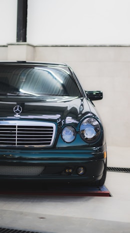 A sleek black Mercedes-Benz car positioned indoors under bright lighting, showcasing its glossy finish and classic front design with a prominent grille and pair of round headlights.