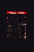 A dimly lit refrigerator filled with various drink bottles is displayed, featuring a prominent red Coca-Cola sign at the top. The contents are mostly obscured by darkness, with only the outlines and some colors of the bottles visible.