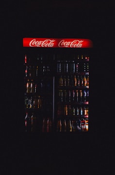 A dimly lit refrigerator filled with various drink bottles is displayed, featuring a prominent red Coca-Cola sign at the top. The contents are mostly obscured by darkness, with only the outlines and some colors of the bottles visible.