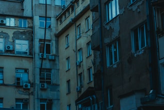 A group of old, worn apartment buildings with peeling paint and visible air conditioning units attached. The structures are multi-storied, with many windows reflecting a cool, dim light. The overall condition of the buildings suggests age and character.