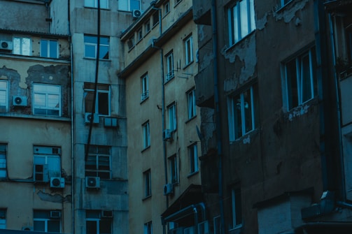 A group of old, worn apartment buildings with peeling paint and visible air conditioning units attached. The structures are multi-storied, with many windows reflecting a cool, dim light. The overall condition of the buildings suggests age and character.