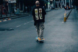 A person wearing a black shirt with graphic prints and beige pants rides a skateboard down an urban street. The scene is slightly blurry, suggesting motion, with a few pedestrians and a car in the distance. There's a yellow and white traffic bollard on the street, and buildings line the roadway under a dim, moody light.