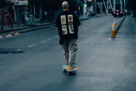 A person wearing a black shirt with graphic prints and beige pants rides a skateboard down an urban street. The scene is slightly blurry, suggesting motion, with a few pedestrians and a car in the distance. There's a yellow and white traffic bollard on the street, and buildings line the roadway under a dim, moody light.