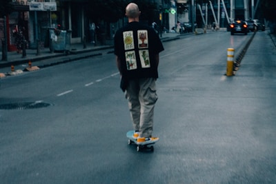 A person wearing a black shirt with graphic prints and beige pants rides a skateboard down an urban street. The scene is slightly blurry, suggesting motion, with a few pedestrians and a car in the distance. There's a yellow and white traffic bollard on the street, and buildings line the roadway under a dim, moody light.