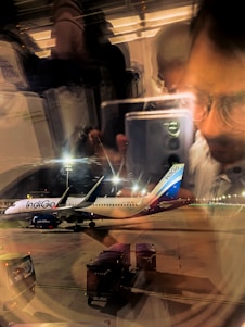 A view from inside an airplane showing a reflection of a person taking a photo with their phone. Outside, an IndiGo airplane is parked at an airport terminal during nighttime, with airport lights illuminating the scene. Baggage carts are visible near the aircraft.