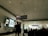 A spacious airport terminal features a digital sign with directions to gates and shopping. A large advertisement with people having their mouths covered is displayed. Silhouettes of travelers, seated and standing, populate the foreground against a backdrop of large windows.