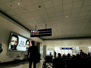 A spacious airport terminal features a digital sign with directions to gates and shopping. A large advertisement with people having their mouths covered is displayed. Silhouettes of travelers, seated and standing, populate the foreground against a backdrop of large windows.