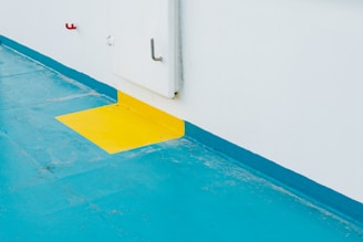 A brightly colored floor features a contrasting yellow marking against a turquoise background adjacent to a white wall. A metal handle is attached vertically on the wall, with a small red hook or object mounted nearby.