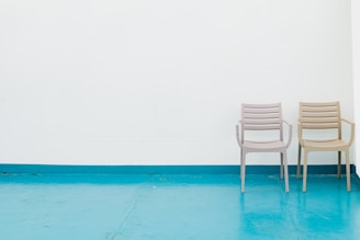 Two empty chairs with armrests are positioned against a plain white wall on a turquoise floor. The chairs are identical in design, featuring horizontal slats, and are positioned side by side slightly apart from each other.