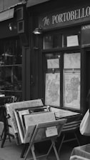 A storefront displaying a collection of vintage maps in wooden frames. The shop window shows maps of the United Kingdom and Europe, with signs indicating they are for sale. The exterior is labeled as 'The Portobello' and features classic details indicative of an antique or historic shop. The scene is black and white, giving it a nostalgic, timeless feel.