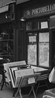 A storefront displaying a collection of vintage maps in wooden frames. The shop window shows maps of the United Kingdom and Europe, with signs indicating they are for sale. The exterior is labeled as 'The Portobello' and features classic details indicative of an antique or historic shop. The scene is black and white, giving it a nostalgic, timeless feel.