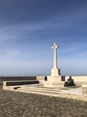 A tall stone cross stands on a raised platform made of stone. The cross is situated in a paved courtyard with a clear blue sky in the background. The cross is the focal point, with tiered steps leading up to it.
