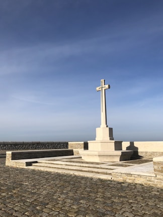 A tall stone cross stands on a raised platform made of stone. The cross is situated in a paved courtyard with a clear blue sky in the background. The cross is the focal point, with tiered steps leading up to it.