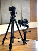 Two professional cameras mounted on tripods are placed on a wooden table. The background consists of a light-colored wall with wooden and stone textures.